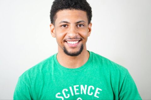 A smiling man wearing a T-shirt stands in a room.